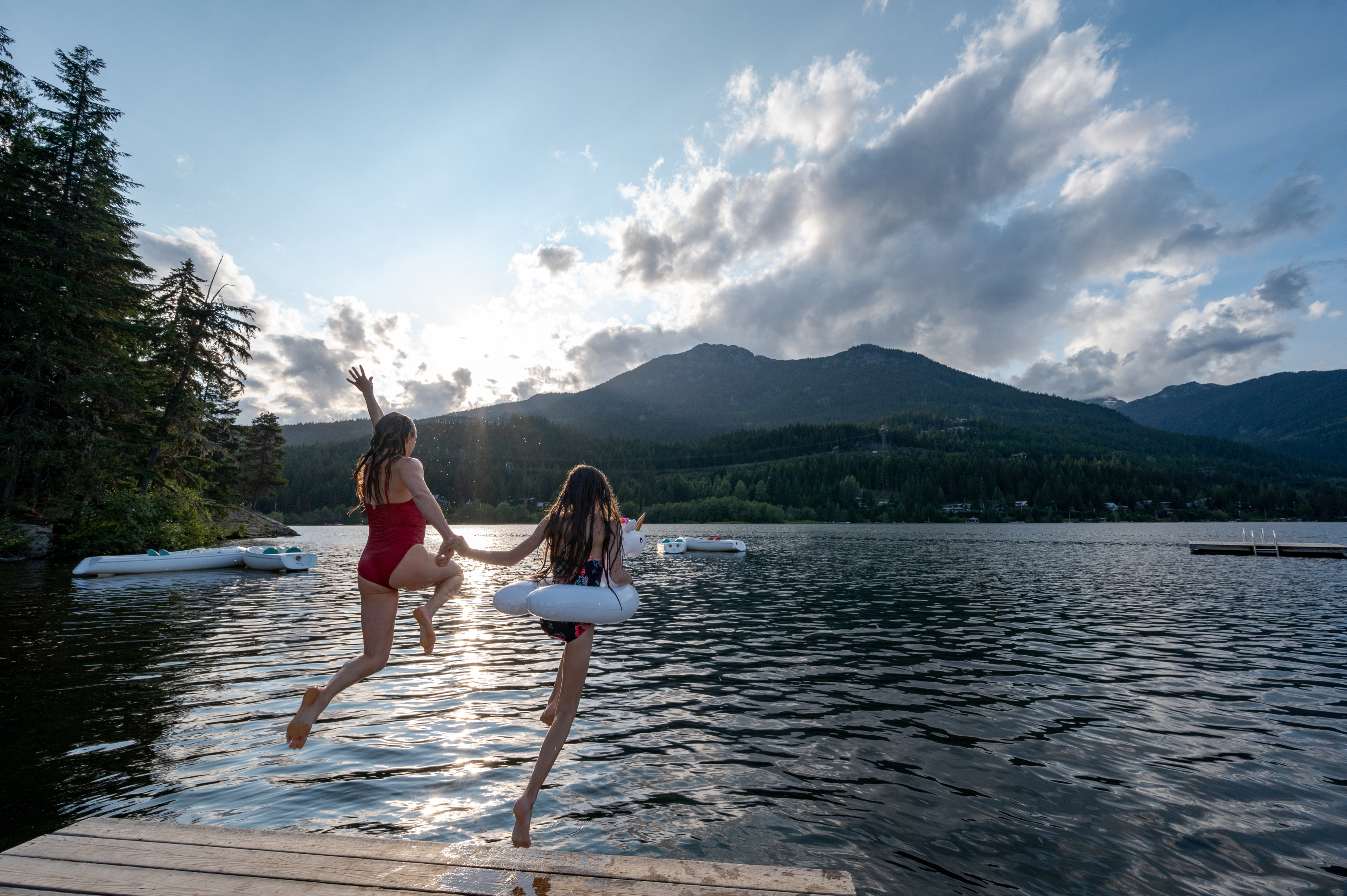 Two people jumping off a dock into a lake with mountains in the background