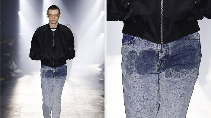 Model on runway in black jacket and faded jeans with wet stain in groin. Close-up shows focus on urine-like stain on denim.