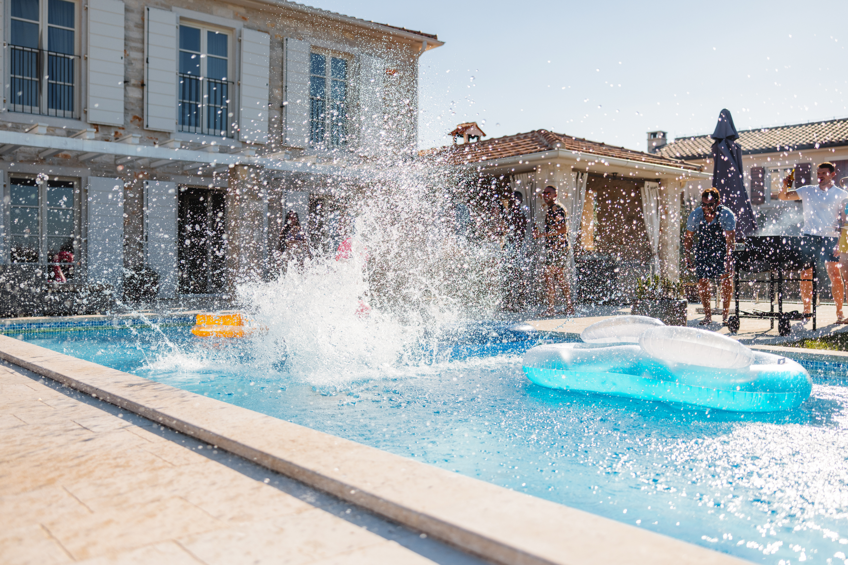 Person splashing into a pool near a pool float, with onlookers by a house in the background