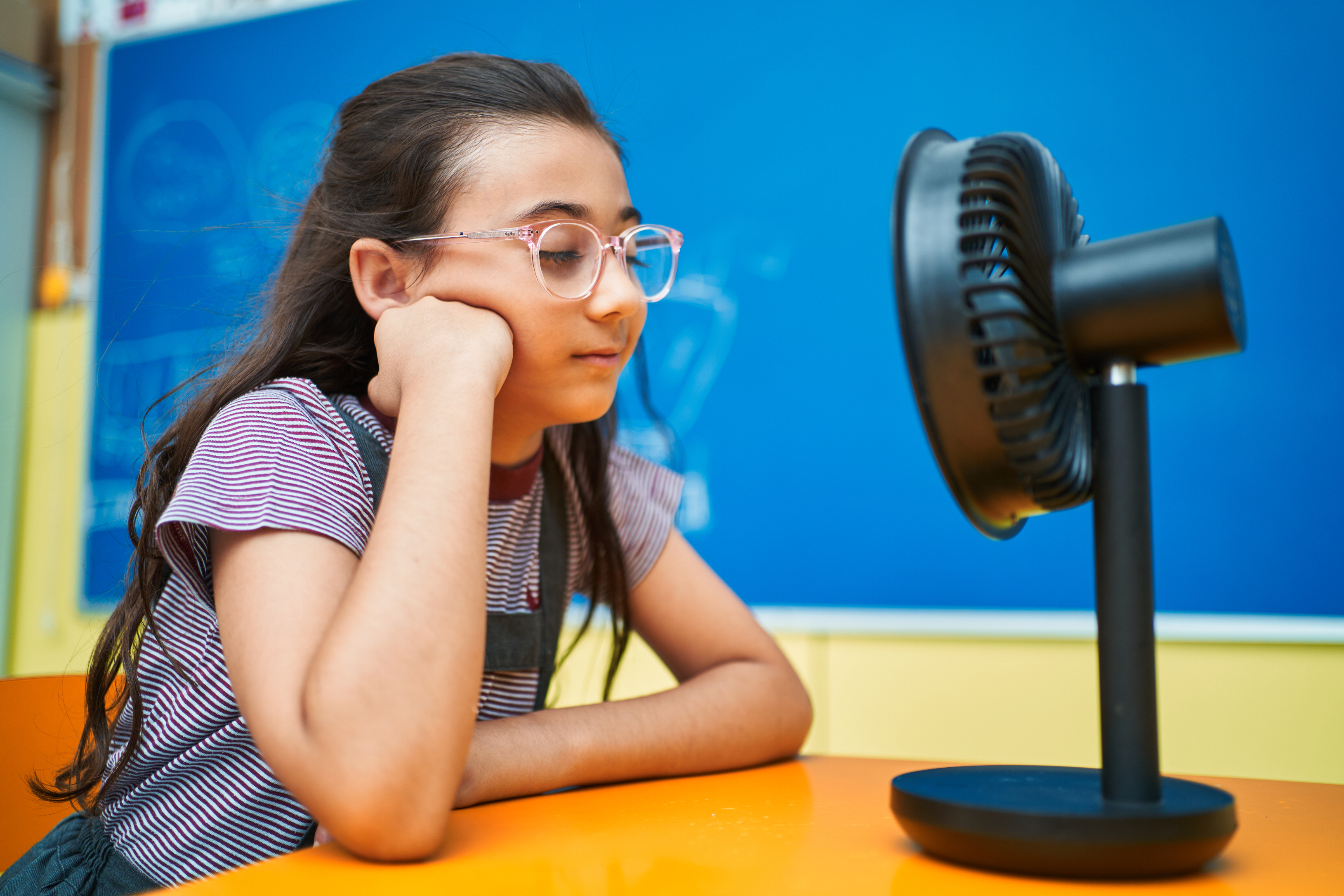 Girl with glasses leans on table, looking at a stationary fan in a classroom environment
