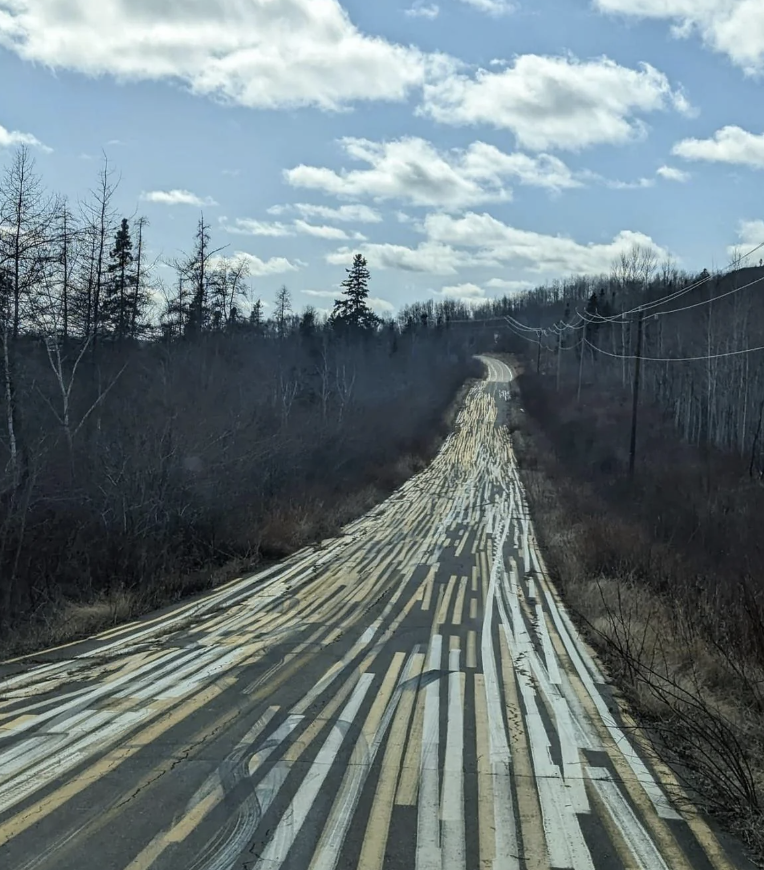 A long straight road with tire tracks and shadows from trees on its surface, under a cloudy sky. No persons