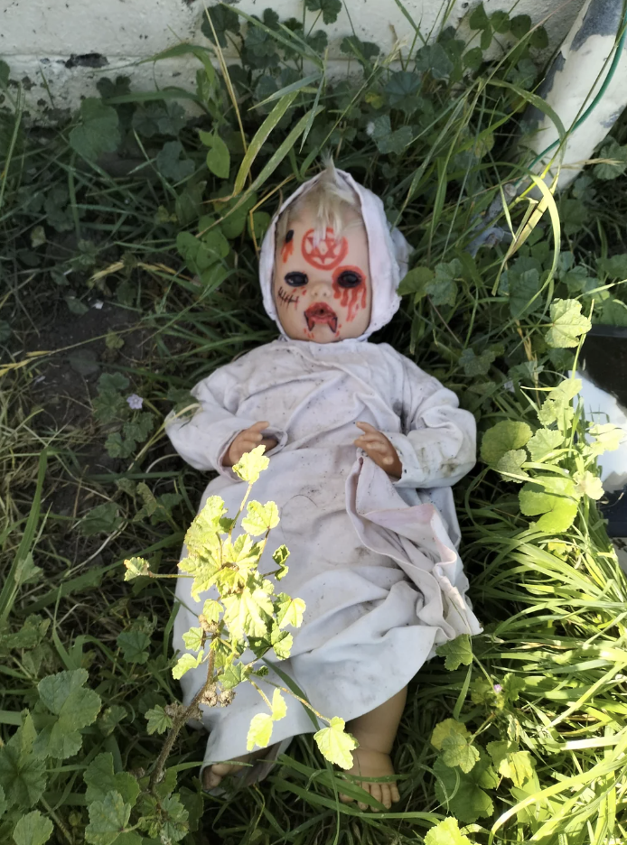 Doll with painted face lying on grass beside a wall, wearing a white outfit