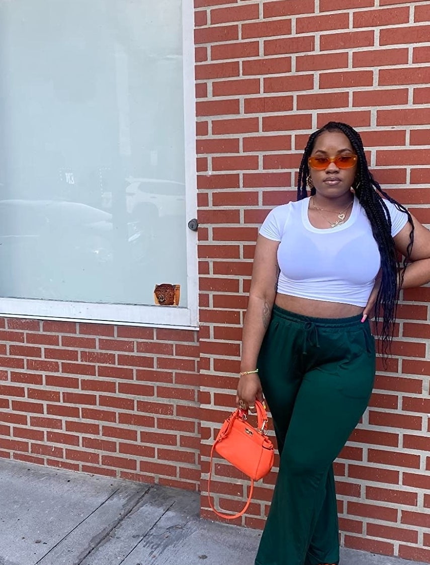 a reviewer in white crop top and green pants with orange bag and shoes posing by a brick wall