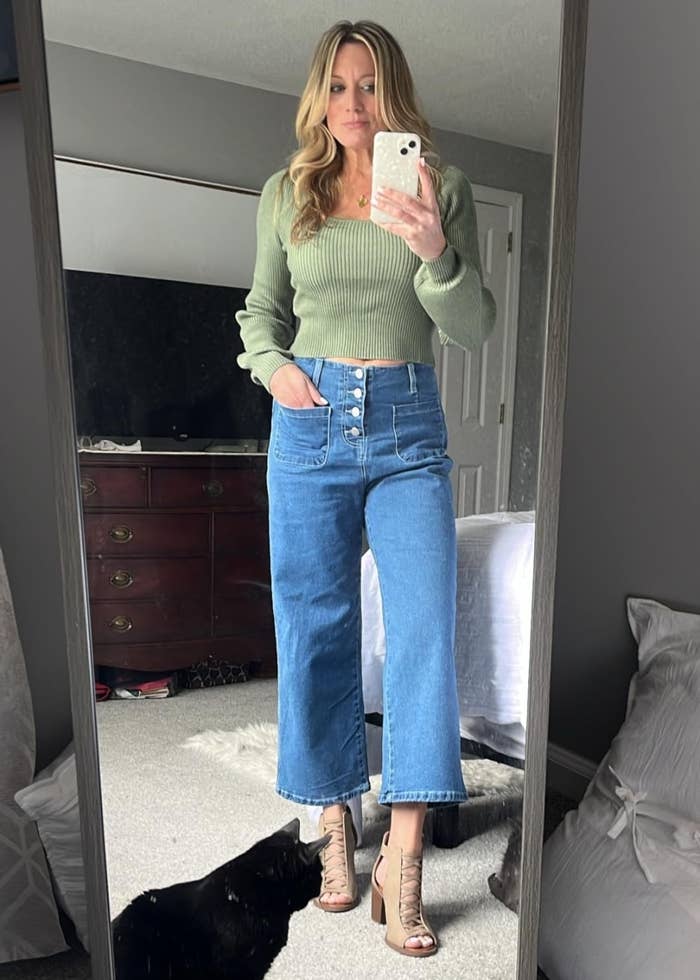 a reviewer in a mirror selfie with a black cat, wearing a green sweater, wide-leg jeans, and heeled sandals