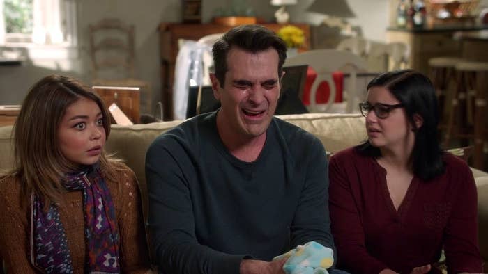 Three people sitting on a couch with surprised expressions, one holding a plush toy. They are TV characters