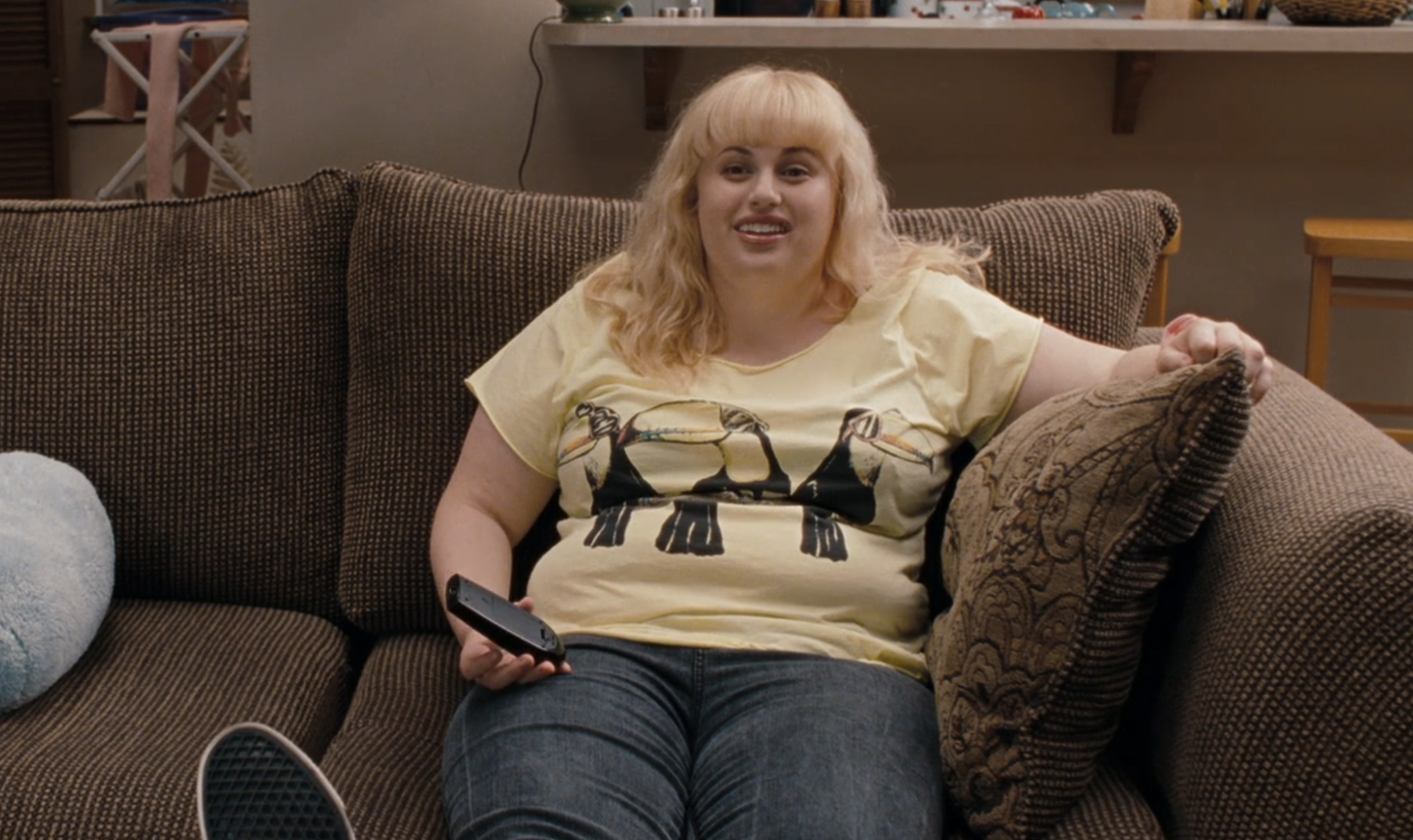A woman sitting on a sofa, holding a remote, wearing a T-shirt with a graphic design, and expressing contentment