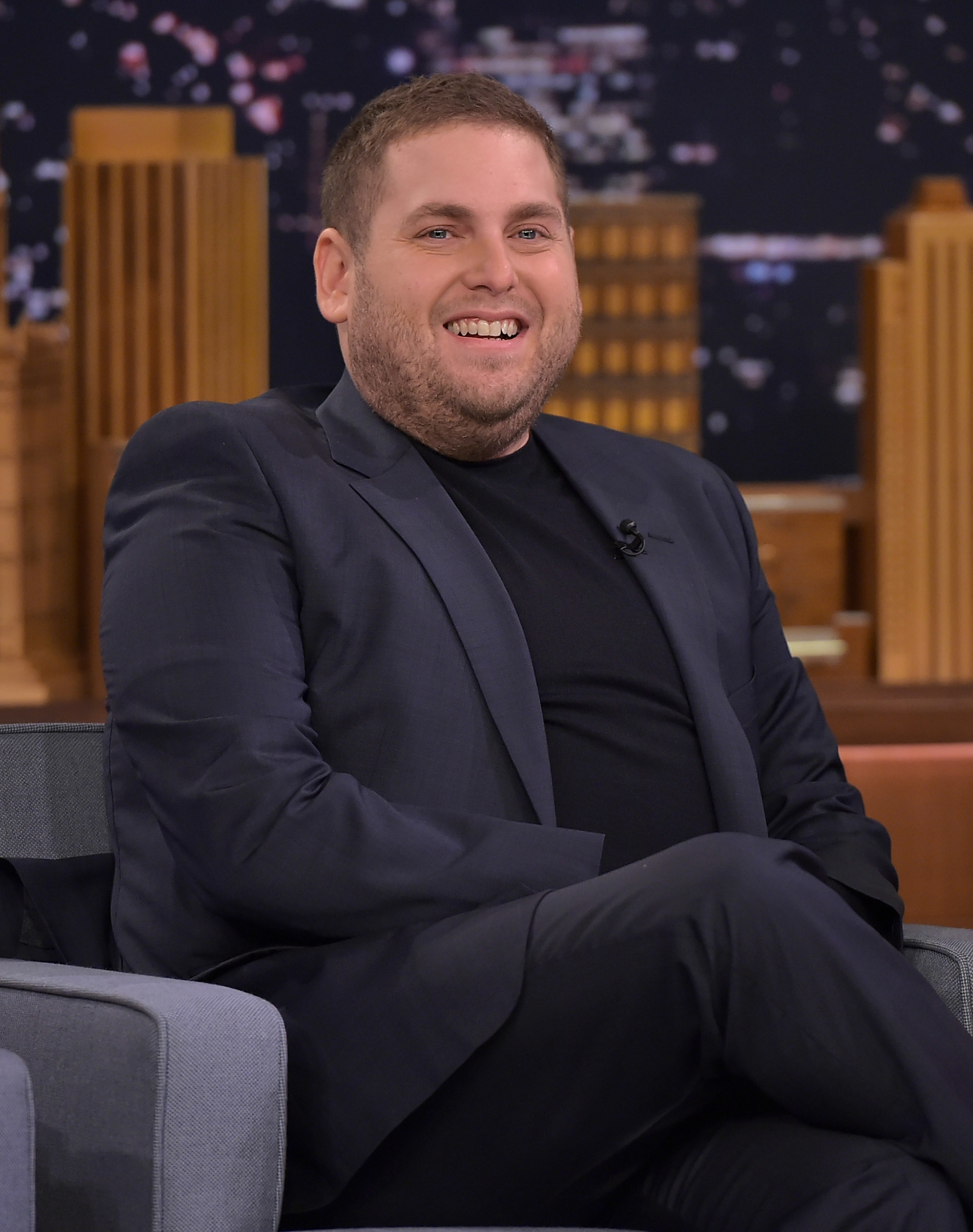 Jonah Hill sits smiling in a suit on a talk show set