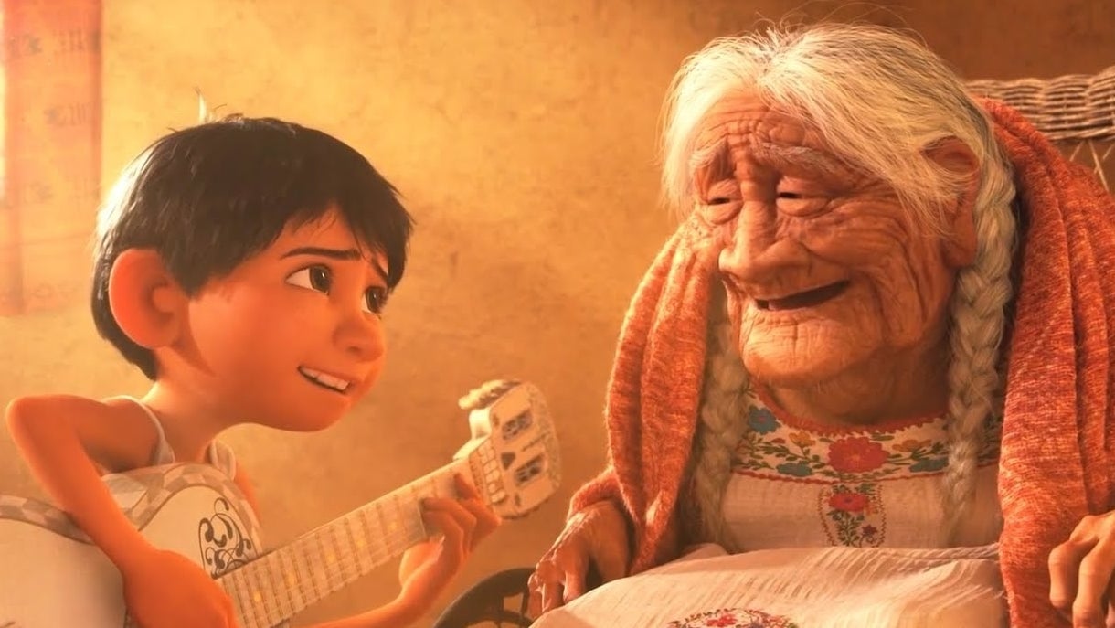 Miguel from Coco smiles at his great-grandmother Coco as he holds a guitar. They are indoors with warm lighting