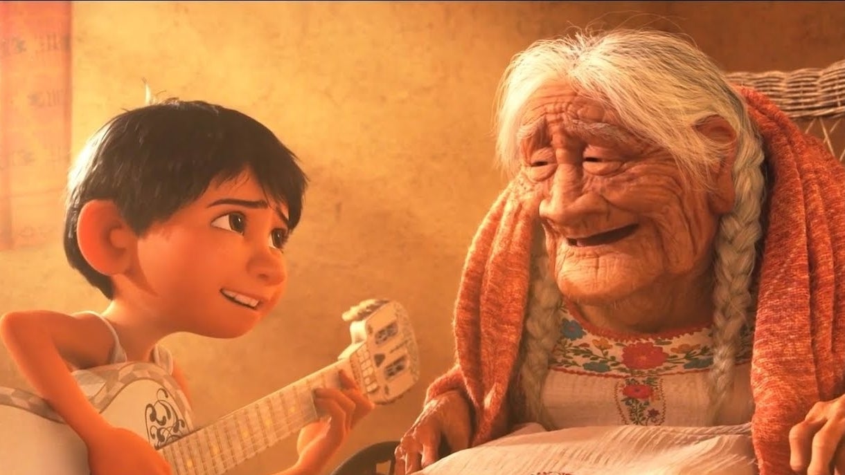 Miguel from Coco smiles at his great-grandmother Coco as he holds a guitar. They are indoors with warm lighting
