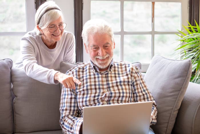 Two elderly individuals smiling, one pointing at a laptop screen, seated on a sofa indoors