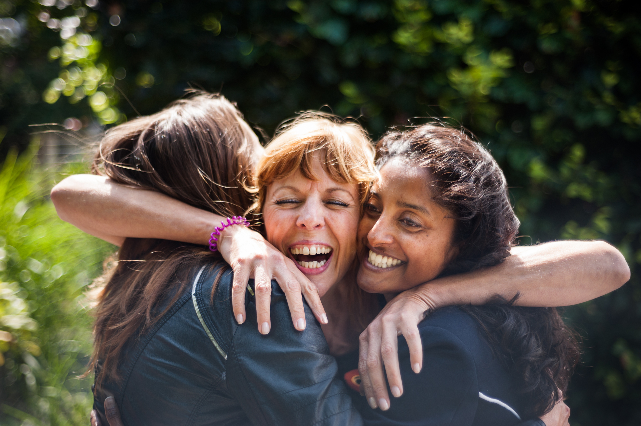Three friends are hugging and smiling outdoors, expressing joy and affection