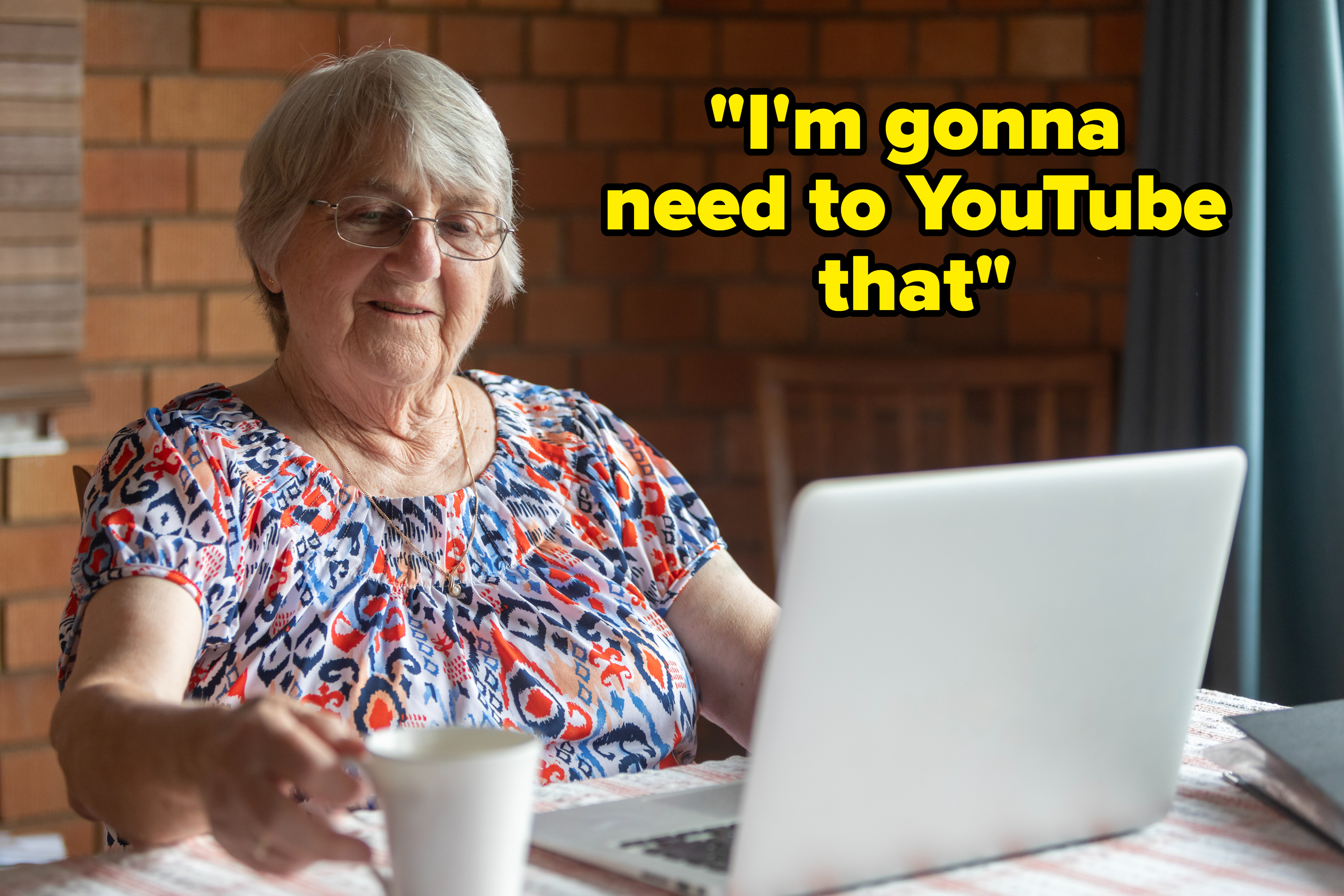 Elderly woman using a laptop at a table with a coffee cup, smiling slightly, in casual printed attire