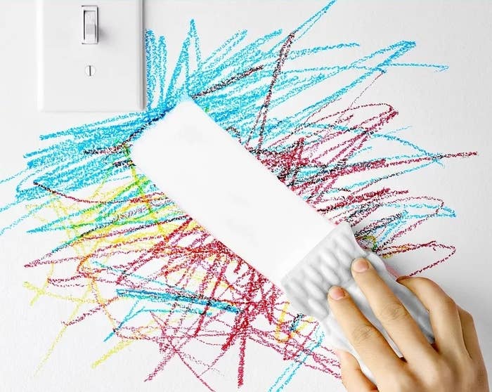 Hand removing crayon marks from a wall with a magic eraser, showcasing a cleaning product