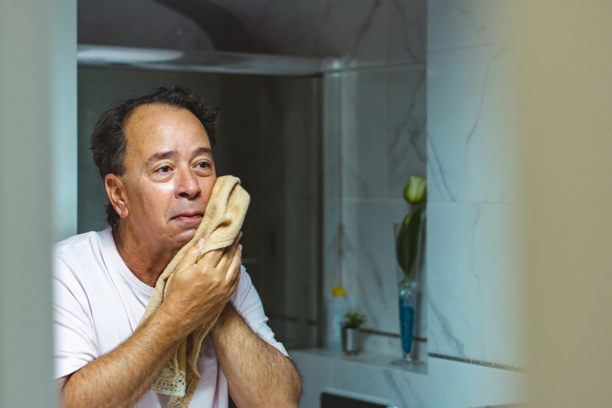 Person in a white shirt wiping their face with a beige towel in front of a mirror