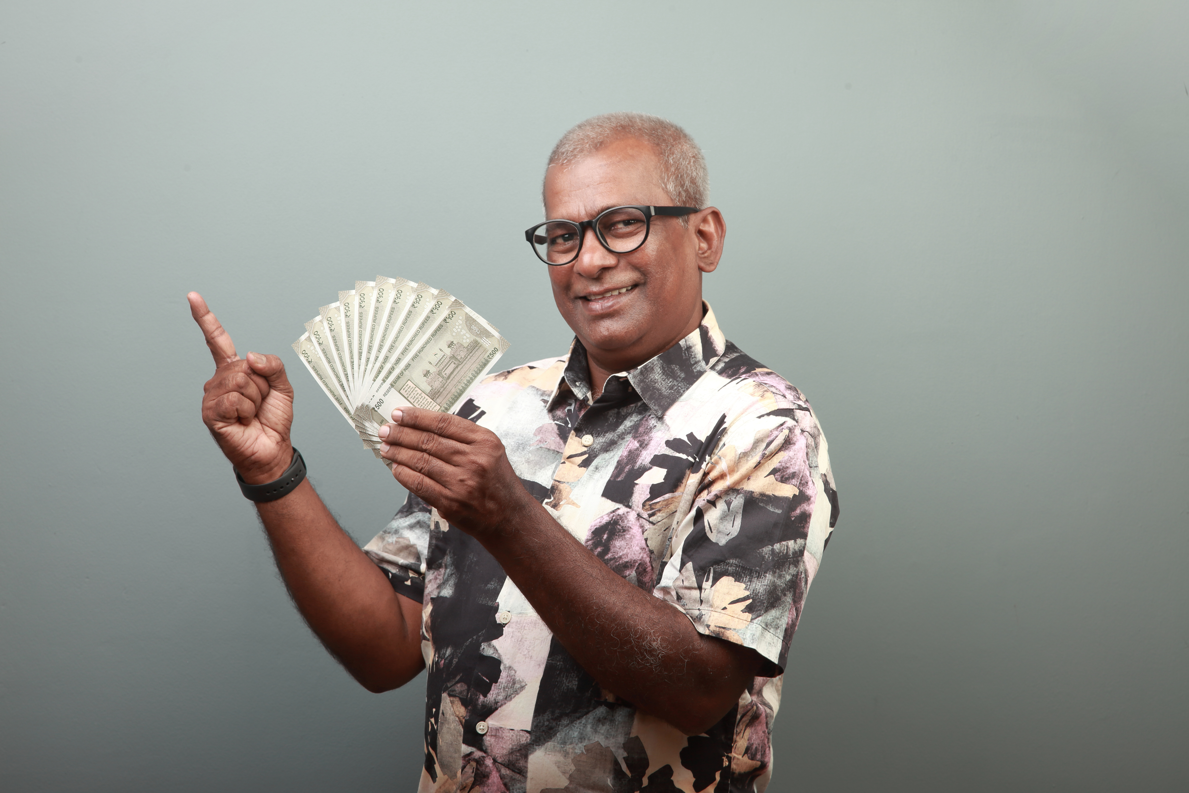 Man in a patterned shirt smiling and pointing to a fan of cash in his hand