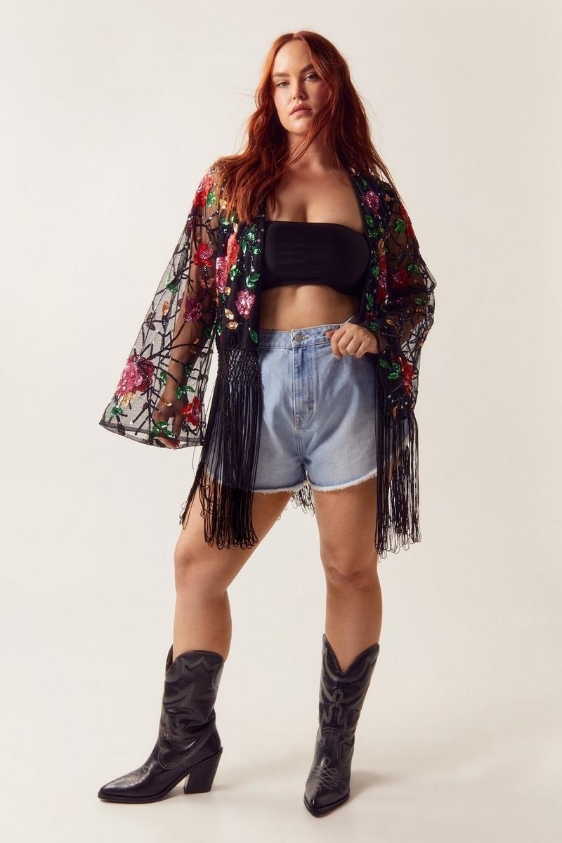 Model poses in a cropped top, denim shorts, fringe jacket, and cowboy boots
