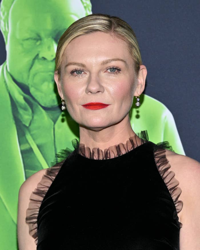 Kirsten Dunst in sleeveless dress with ruffled accents, lipstick, standing before a graphic backdrop