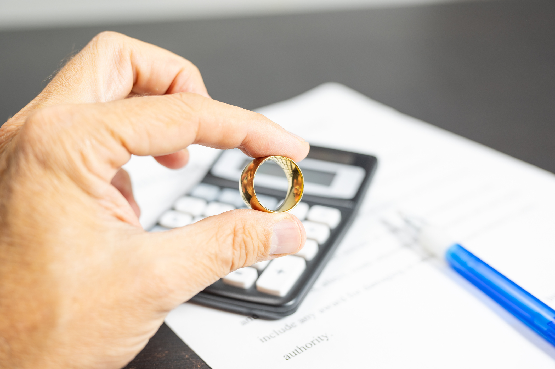 Hand holding a wedding ring above a calculator on a document, symbolizing financial considerations in marriage