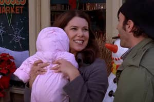Lorelai Gilmore from Gilmore Girls smiles while holding a baby and talking to Luke Danes outside