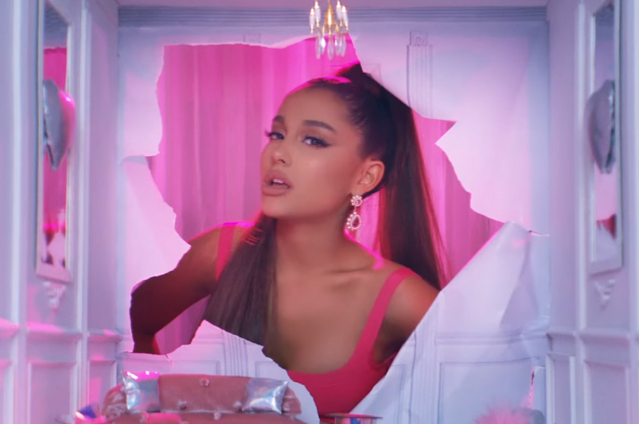 Ariana Grande poses in a pink room with a broken heart-shaped wall behind her