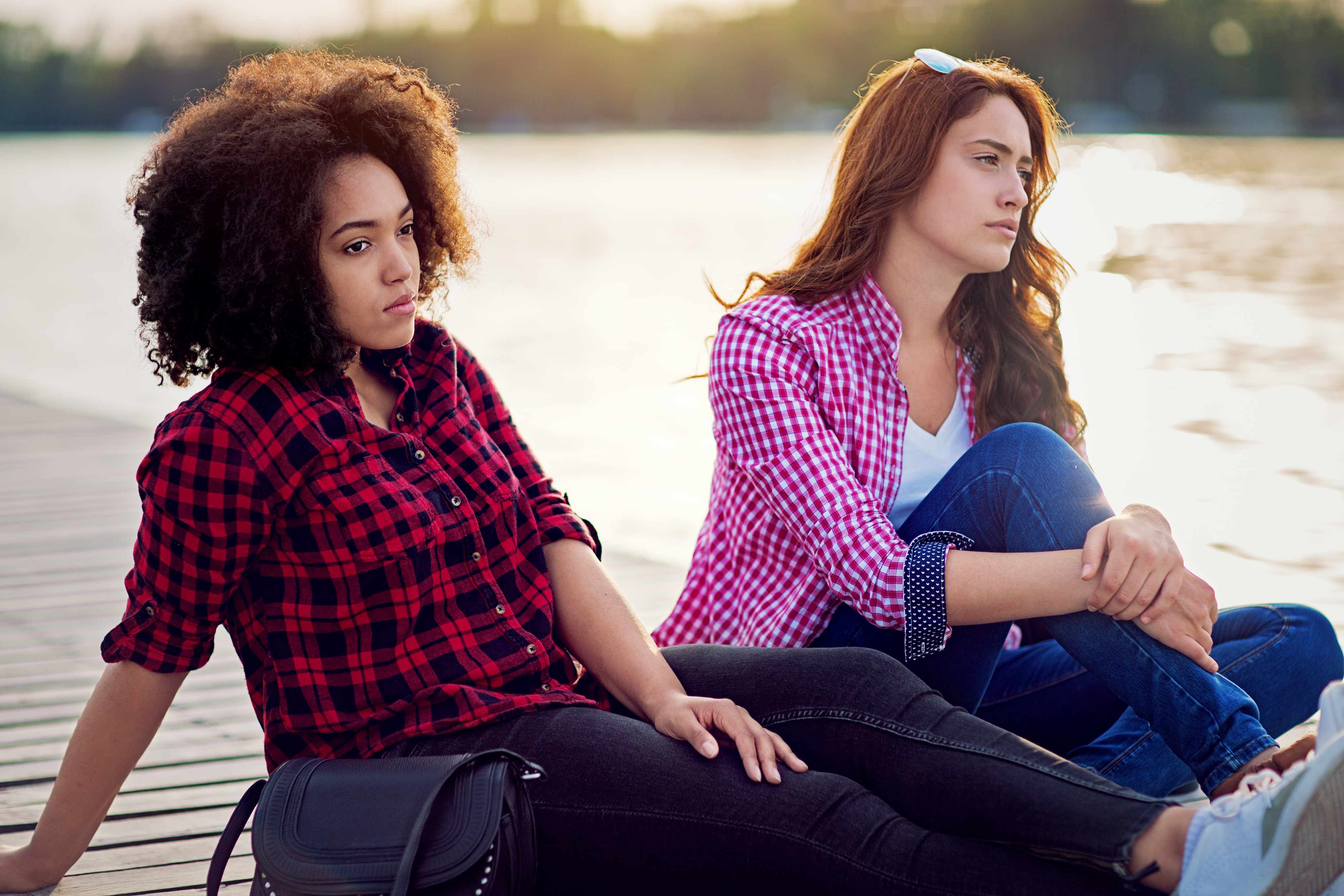Two women sitting by the water, wearing casual plaid shirts and jeans