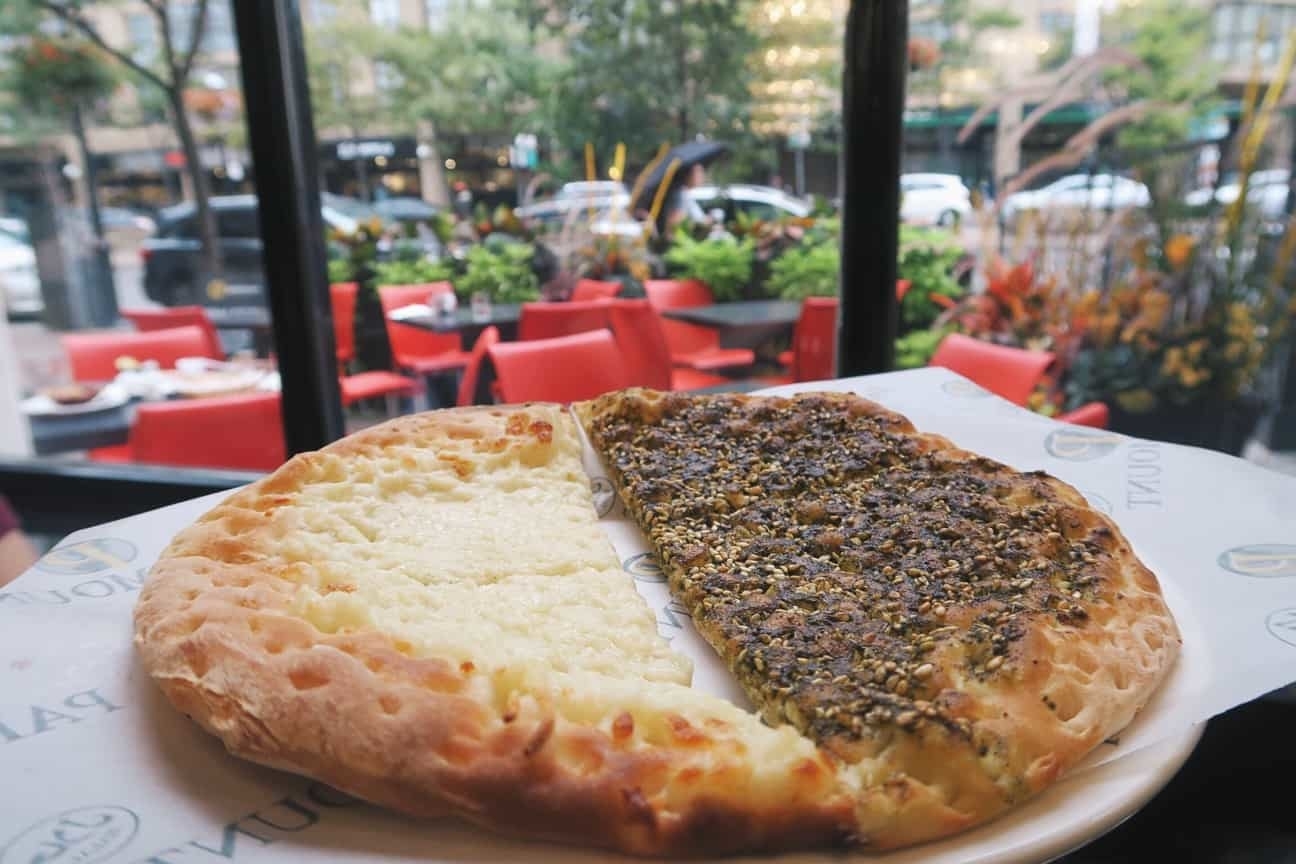 Half cheese, half za&#x27;atar manakish on a plate, viewed from indoors with a street-side cafe in the background
