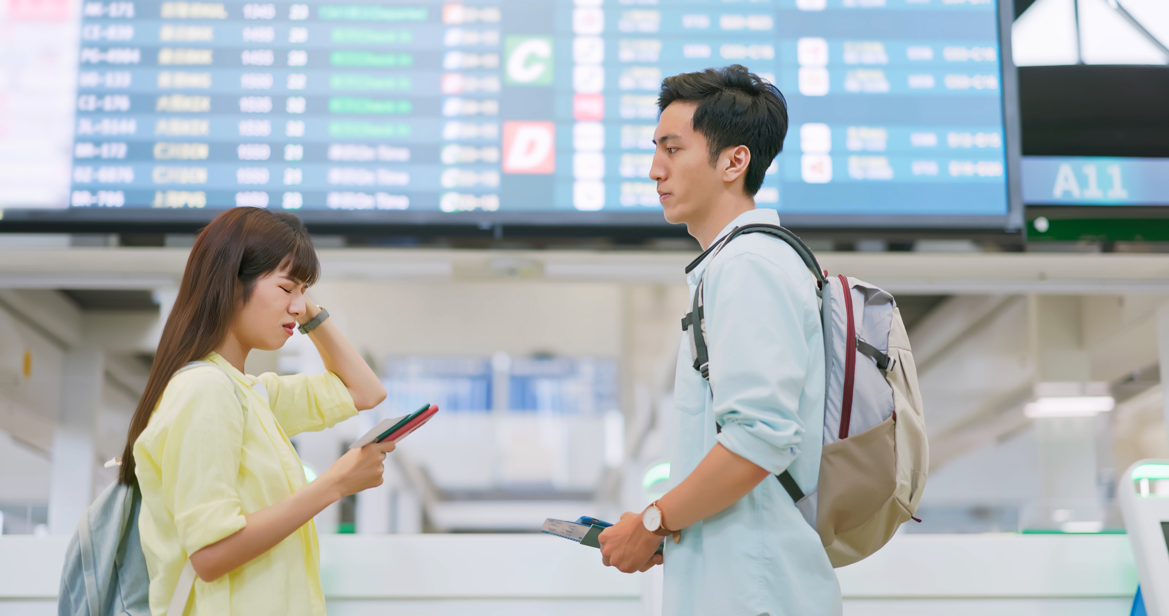 Two people standing in an airport terminal, looking at phone and tickets, with flight information board in the background