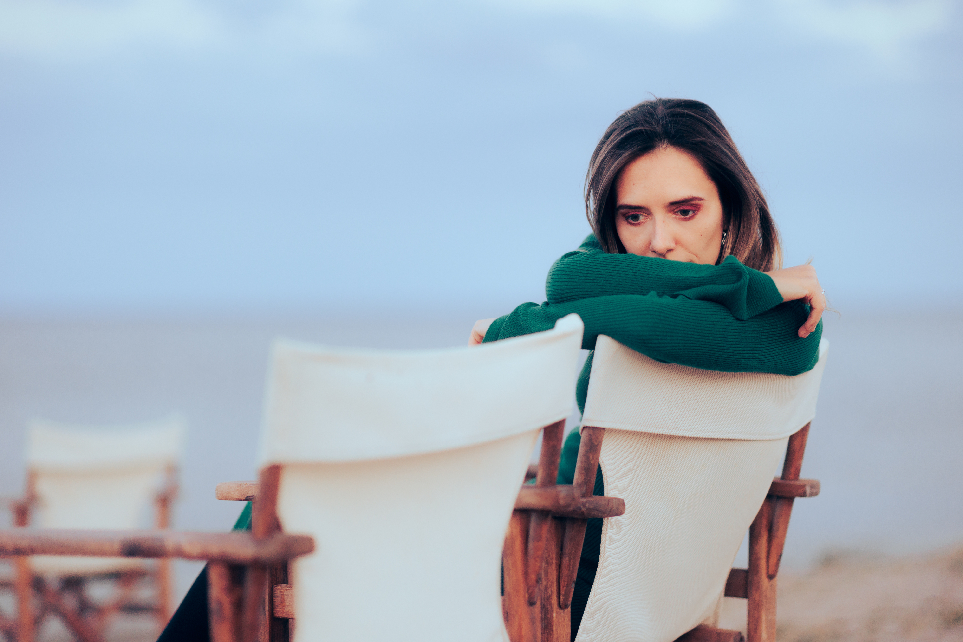 Woman sitting in a deck chair on a beach, wrapped in a sweater, resting her chin on her arms