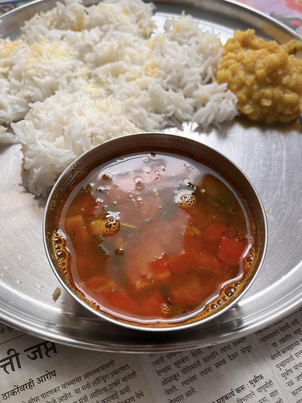 A bowl of clear soup with vegetables beside rice and lentils on a plate, over a newspaper
