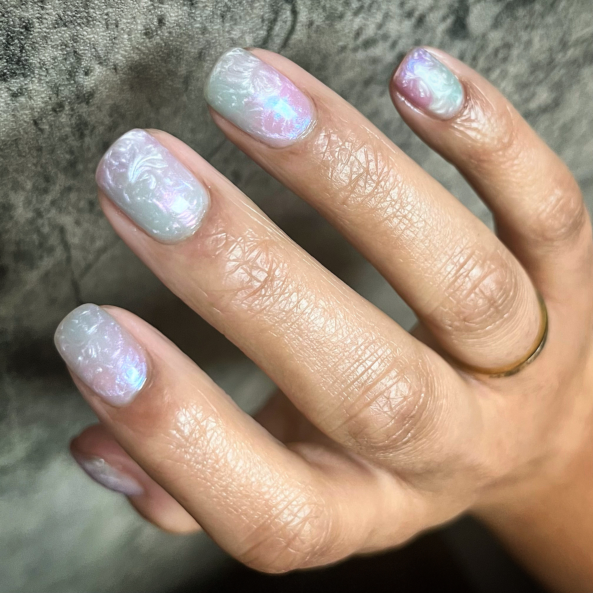 Hand sporting iridescent manicured nails with a subtle sheen, showcasing a ring on the ring finger