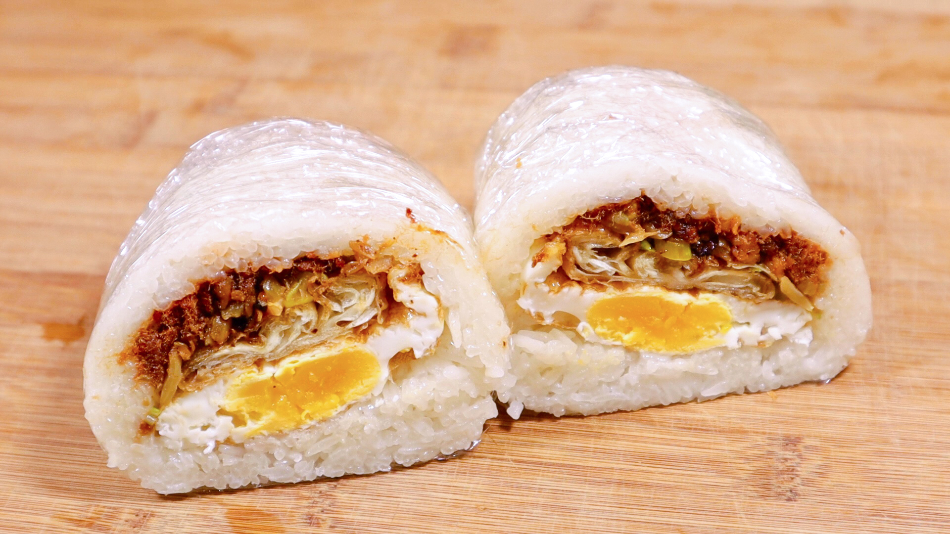 Two halves of a rice ball with meat, egg, and vegetable filling on a wooden surface