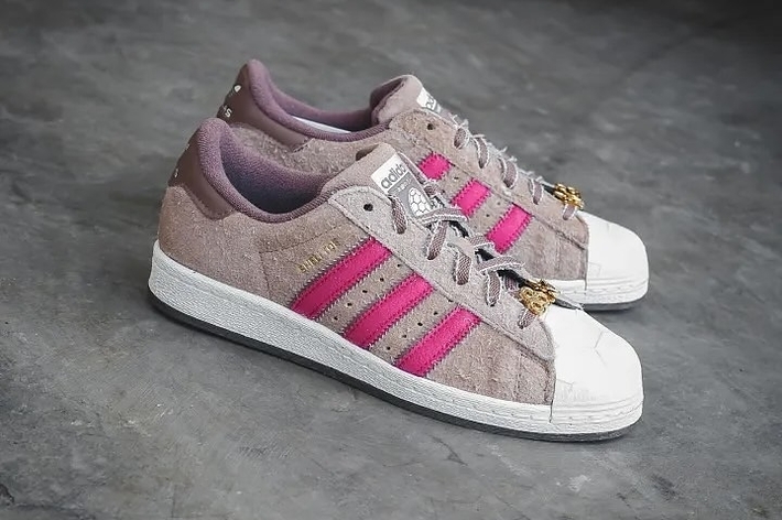 A pair of Adidas sneakers with pink stripes and a gold bee emblem on a concrete surface