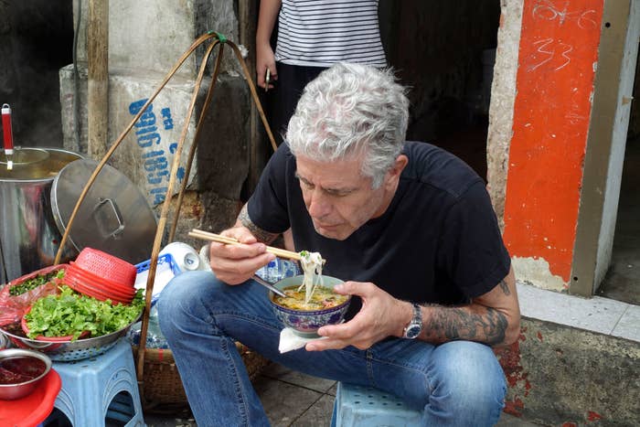 Man sitting on a stool eating noodles from a bowl at a street food stall