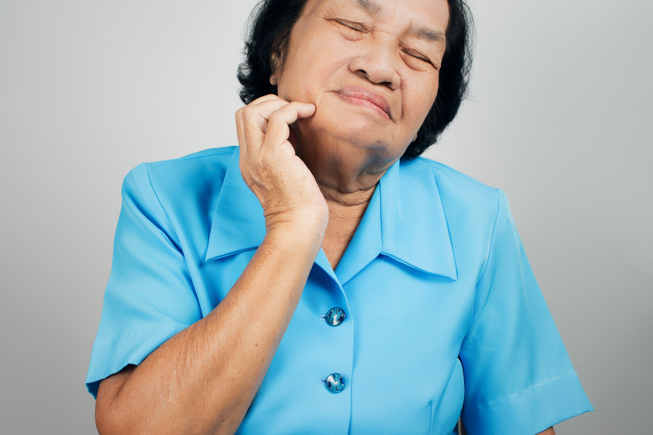 Elderly woman pinching her cheek, eyes closed, with a pained or uncomfortable expression