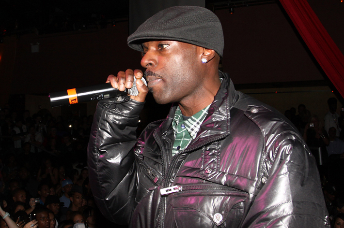 A musician performs onstage holding a microphone, wearing a trendy jacket and cap