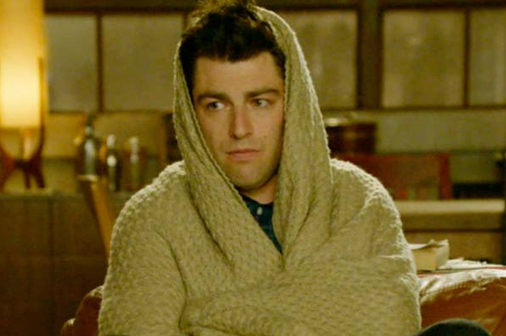 Person wrapped in a beige blanket looking tired, in a room. Character from a TV show or movie