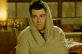 Person wrapped in a beige blanket looking tired, in a room. Character from a TV show or movie