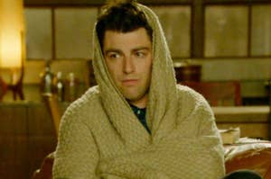 Person wrapped in a beige blanket looking tired, in a room. Character from a TV show or movie