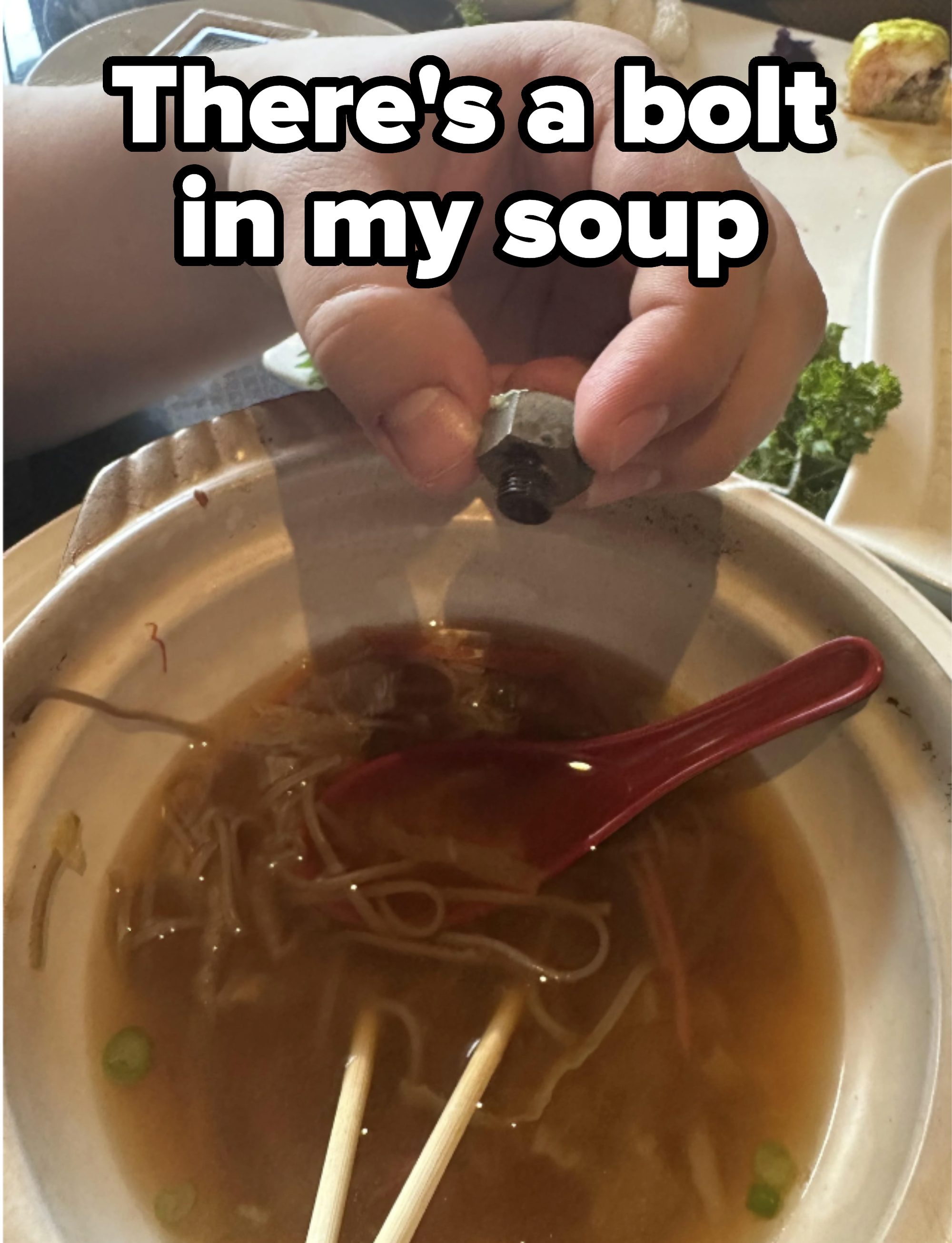 Hand holding a small object over a bowl of noodle soup with chopsticks and a spoon