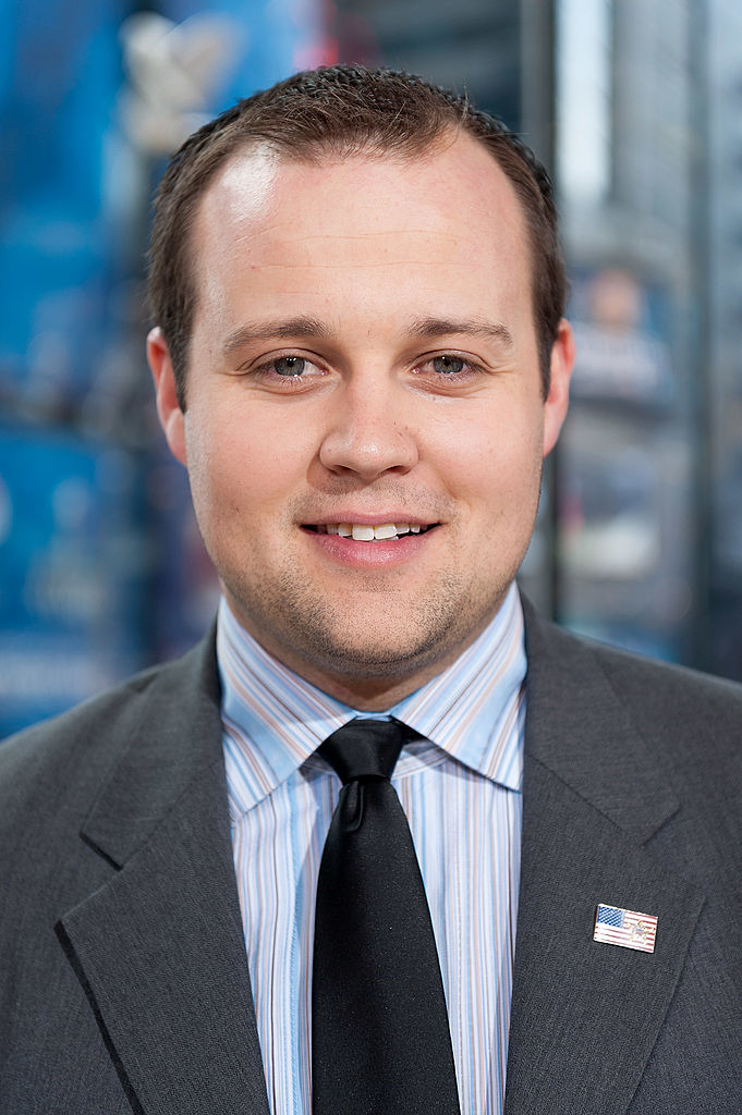 Josh Duggar in a suit with striped tie and American flag lapel pin smiling at the camera