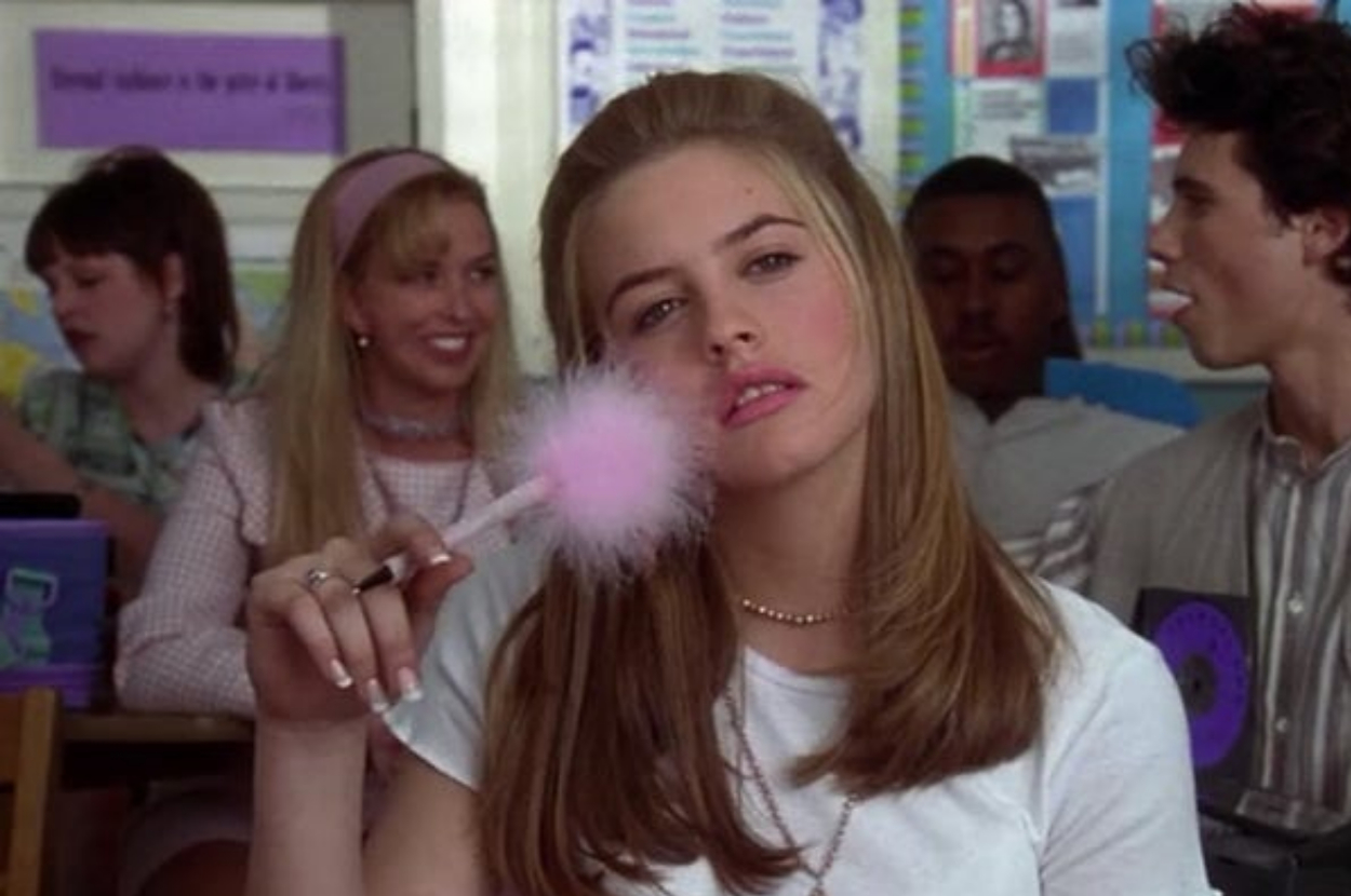 Cher Horowitz, a character from "Clueless," sits in a classroom holding a pink feather pen