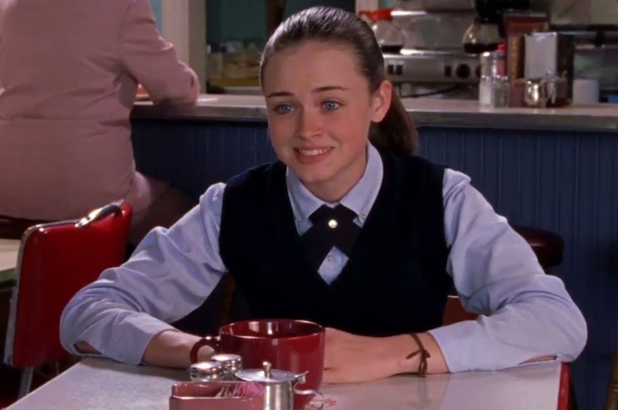 Rory Gilmore sits at a diner booth wearing a sweater and collared shirt