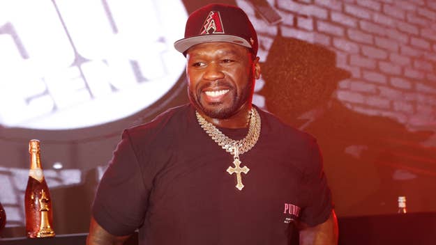 Man smiling at an event with microphone and branded backdrop, wearing a cap, T-shirt, and chain necklace
