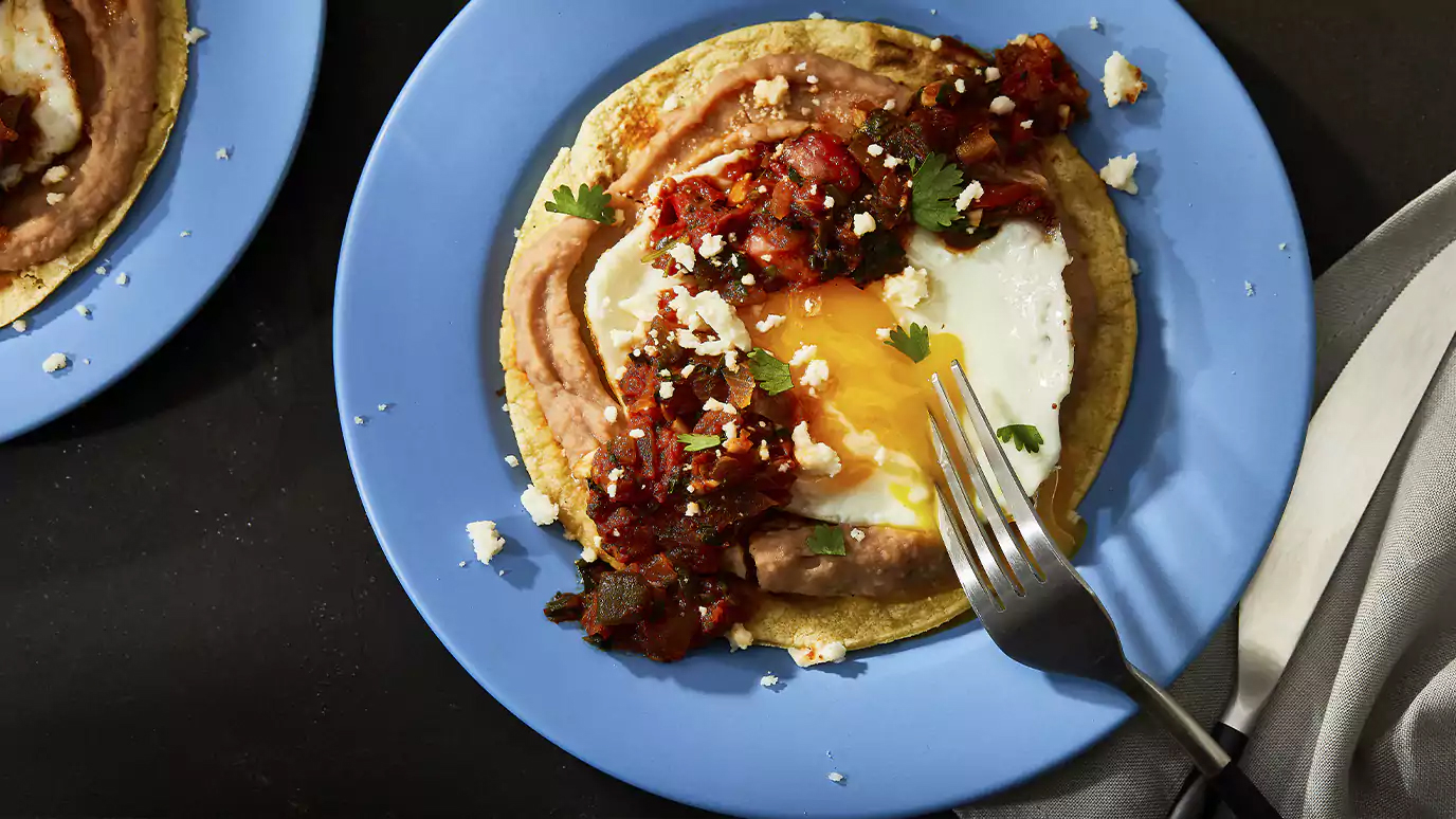 Fried egg on a tortilla with toppings on a blue plate, a fork cutting into the yolk
