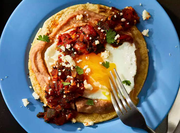 Fried egg on a tortilla with toppings on a blue plate, a fork cutting into the yolk