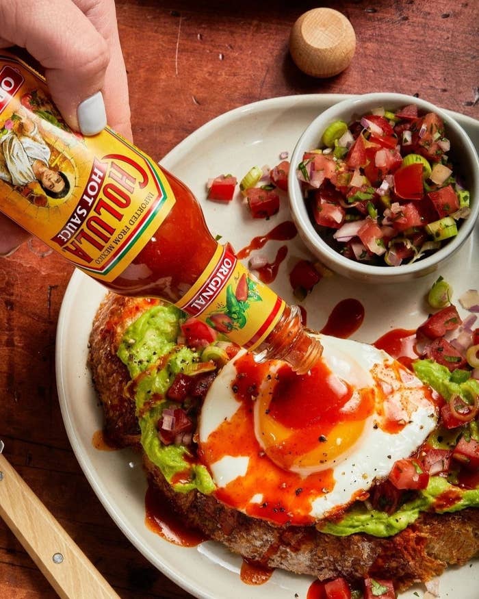 A hand pouring hot sauce on an egg-topped avocado toast with a side of pico de gallo