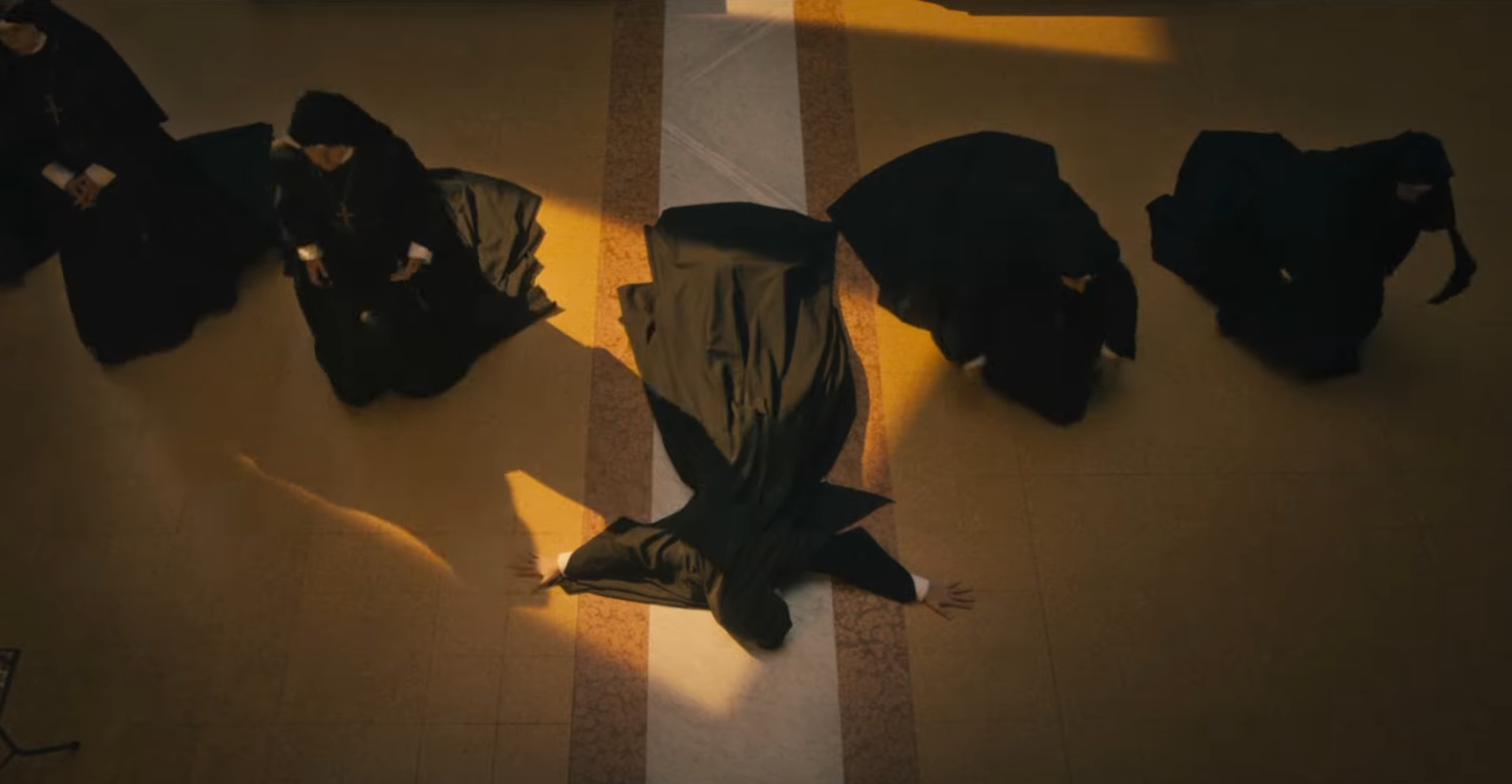 Four people in dark attire lying on the floor around a bright line, creating a dramatic shadow pattern