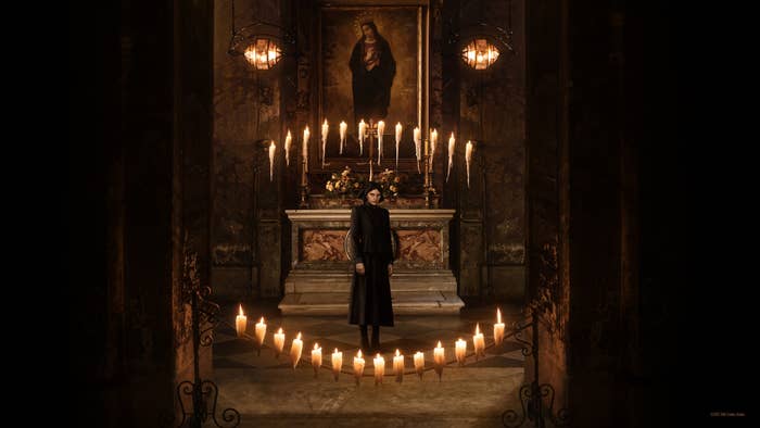 Person in a long coat standing in a dimly lit room with candles and a portrait on the wall