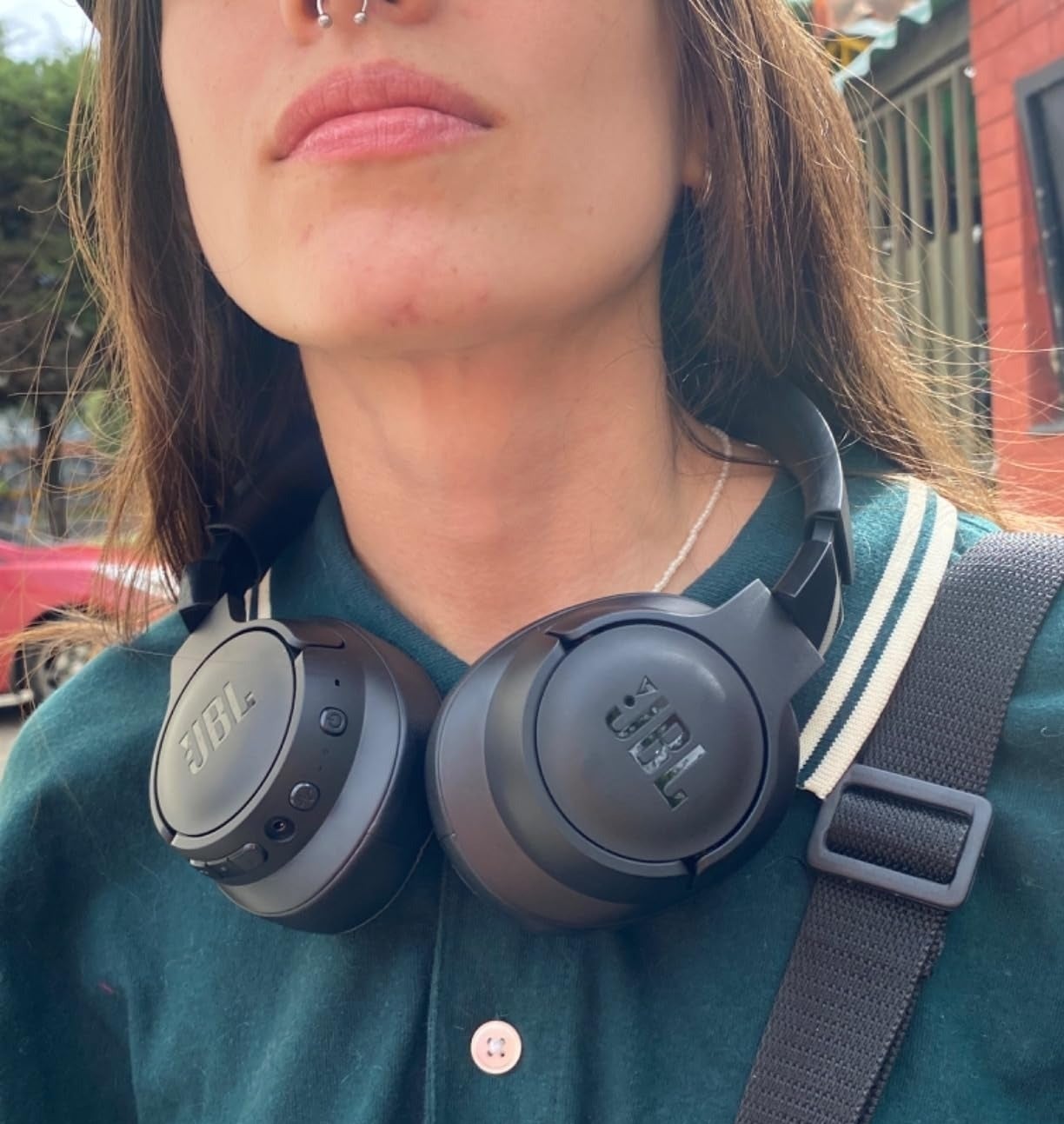 a reviewer wearing the black JBL headphones around their neck with a button-up shirt and a bag strap