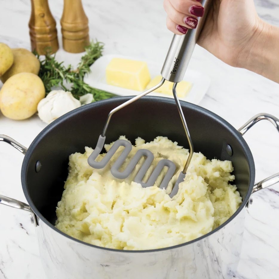 A person&#x27;s hand holding a potato masher over a pot filled with mashed potatoes